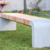 Moderne Sitzbank mit abgerundeten Füßen aus Beton und längs verlaufenden Holzlatten steht in einer öffentlichen Grünanlage.
