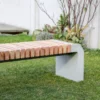 Moderne Sitzbank mit abgerundeten Füßen aus Beton und längs verlaufenden Holzlatten steht in einer öffentlichen Grünanlage.