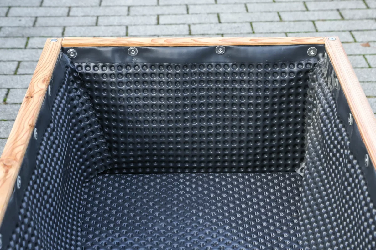 Pflanzkübel aus Lärchenholz, ausgekleidet mit Noppenbahn, fahrbar durch Schwerlastrollen an Unterseite, steht auf öffentlichem Platz.