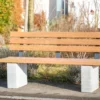 Rollstuhlgerechte Sitzbank aus Hartholz und nach innen versetzten Standfüßen aus Beton steht auf einer öffentlichen Fläche mit Begrünung im Hintergrund