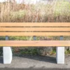 Rollstuhlgerechte Sitzbank aus Hartholz und nach innen versetzten Standfüßen aus Beton steht auf einer öffentlichen Fläche mit Begrünung im Hintergrund