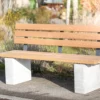 Rollstuhlgerechte Sitzbank aus Hartholz und nach innen versetzten Standfüßen aus Beton steht auf einer öffentlichen Fläche mit Begrünung im Hintergrund