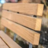 Rollstuhlgerechte Sitzbank aus Hartholz und nach innen versetzten Standfüßen aus Beton steht auf einer öffentlichen Fläche mit Begrünung im Hintergrund